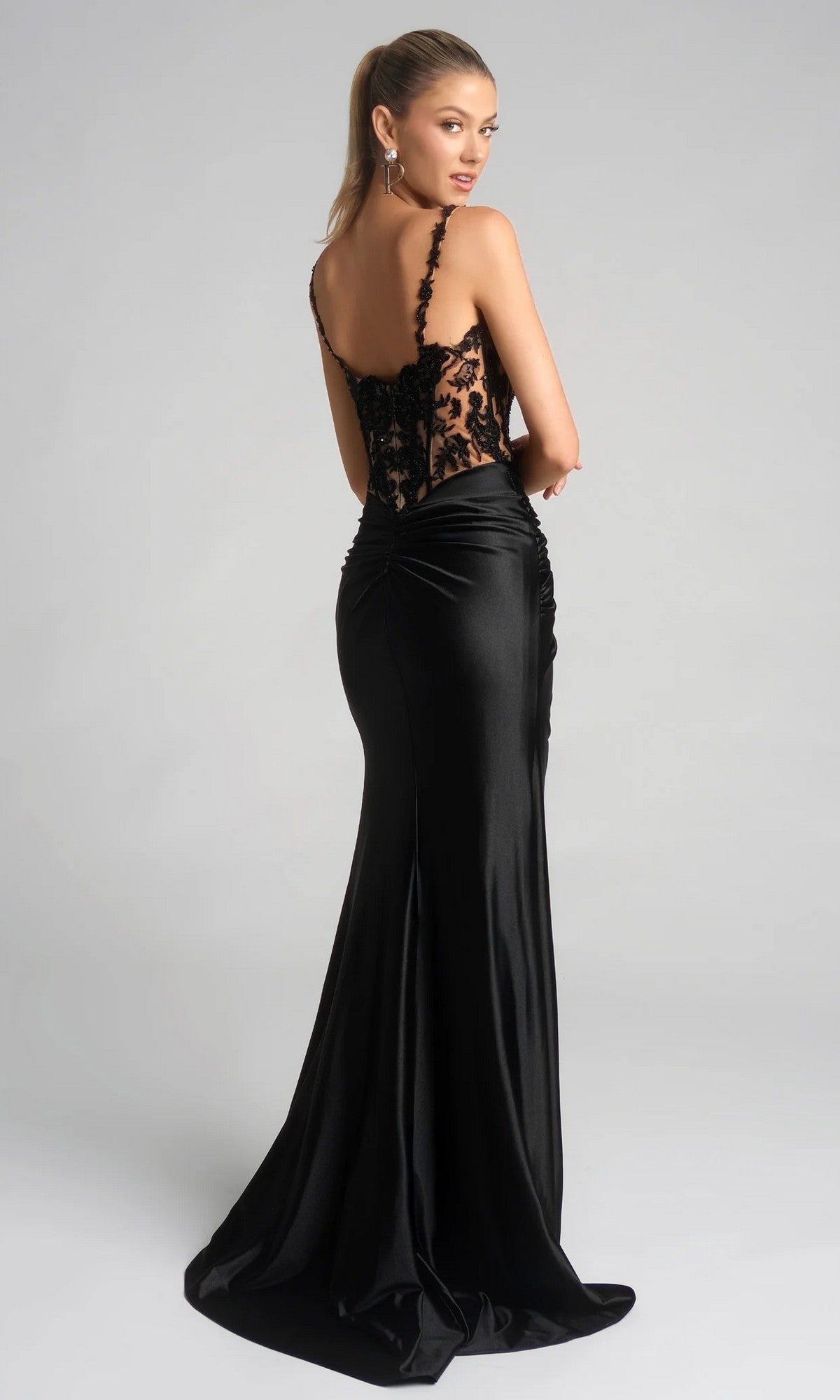 Woman in the Portia and Scarlett XO119 long formal dress, featuring a black lace embroidered corset and satin skirt, stands looking over her shoulder against a plain gray background.